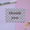Kaart met een roze en oranje geblokt patroon met de tekst 'Thank You' en een illustratie van een lachend gezichtje. Bedoelt om iemand te bedanken.