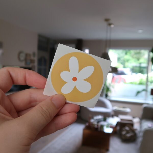 Een gele ronde sticker met een witte bloem
