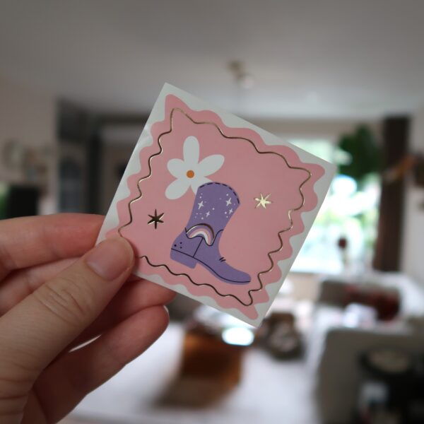 Roze vierkante sticker met een lila cowboy laars.
