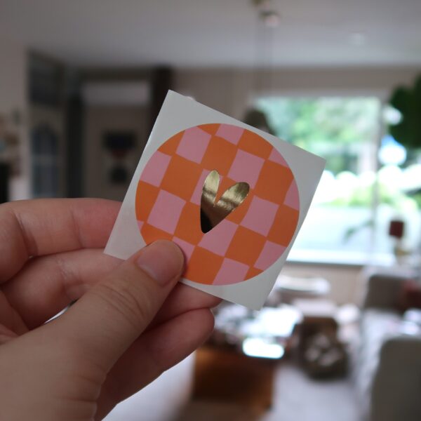 Ronde sticker met oranje en roze geblokt grid achtergrond en een gouden hartje.