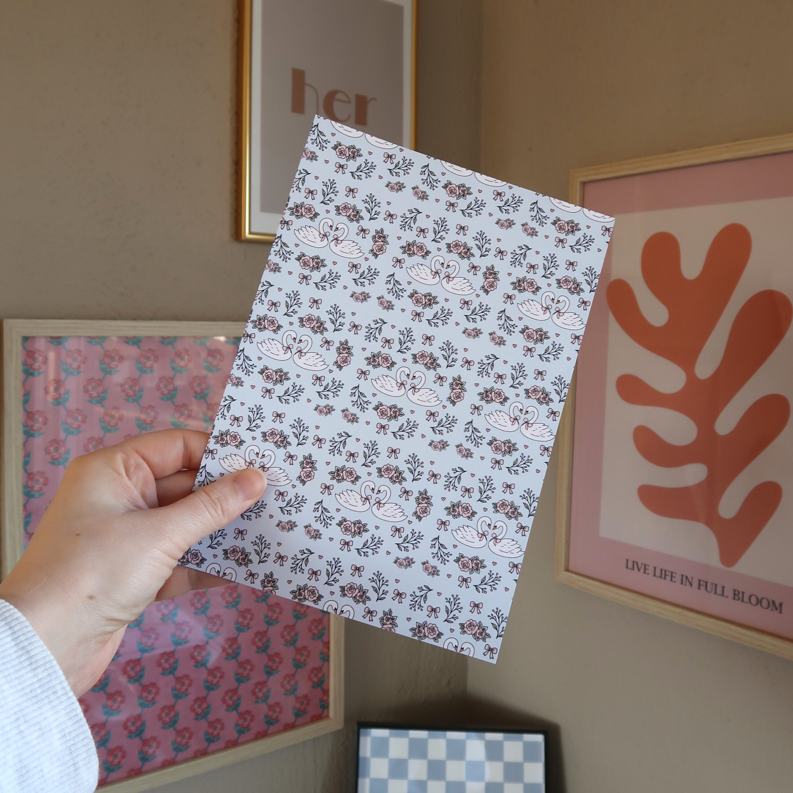 Een hand die een vel papier vast houdt met een illustratie van zwanen, strikjes, rozen, hartjes in pasteltinten licht blauw en roze