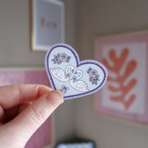 Hand die een losse sticker vast houdt in de vorm van een hartje met de illustratie van twee zwanen, in pastel tinten.
