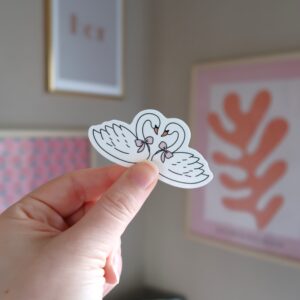 Hand die een losse sticker vast houdt met een illustratie van twee verliefde zwanen