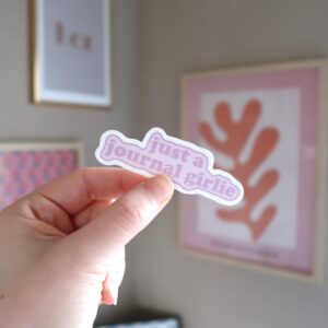Een hand die een losse sticker vast houdt met de tekst just a journal girlie.